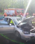 Uszkodzony samochód osobowy. Wokół stoją strażacy, ratownicy medyczni. W tle samochód gaśniczy i ambulans