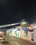 Szereg budynków, znad jednego unosi się dym. Rozstawiona jest drabina mechaniczna nad dachem budynku. Strażacy są na dachu i podają wodę.