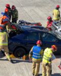 Dwa wraki samochodów leżą na betonowym placu. Wokół aut ustawieni są strażacy ubrania specjalne. Z boku stoi