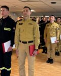 Pododdział strażaków w mundurach stoi na zbiórce w pomieszczeniu. Przed frontem szyku stoi dwóch wyróżnionych strażaków