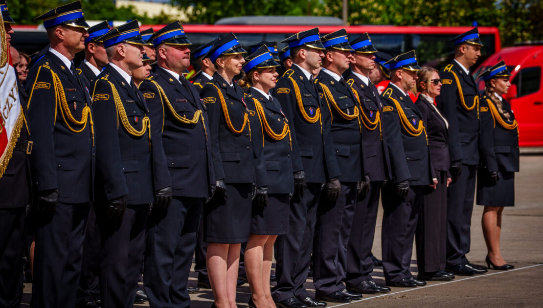Pododdział kilkudziesięciu strażaków w mundurach wyjściowych stoją w szeregu na placu. W tle widać fragmenty czerwonych pojazdów i drzewa.