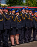 Pododdział kilkudziesięciu strażaków w mundurach wyjściowych stoją w szeregu na placu. W tle widać fragmenty czerwonych pojazdów i drzewa.