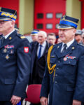 Zastępca komendanta głównego Państwowej straży pożarnej oraz komendant Szkoły Podoficerskiej stoją w ubraniach wyjściowych. Za nimi w tle widać ludzi i czerwone bramy garażowe