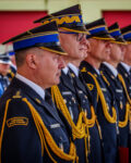 W szeregu stoi 9 komendantów Państwowej Straży Pożarnej. Są w ubraniach wyjściowych. Za nimi w tle widać kilka osób.