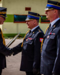 Zastępca komendanta głównego PSP podaje rękę komendantowi Szkoły Podoficerskiej. Po lewej stronie komendanta szkoły stoi dowódca JRG. Wszyscy są w ubraniach wyjściowych. W tle widać sztandar i elewację budynku.