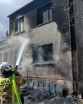 Strażak trzyma w ręku wąż gaśniczy i podaje wodę na okopconą ścianę budynku. Na ziemi widać tlące się resztki jakiegoś materiału.