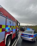 na Jezdni z lewej strony widać kawałek auta strażackiego. Po prawej stoi auto policji. Z tyłu w tle widać uszkodzone auto osobowe i strażaków.