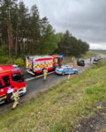 Na jezdni stoja dwa auta strażackie i jedno auto policji. Przed nimi w poprzek drogi stoi uszkodzone auto osobowe.