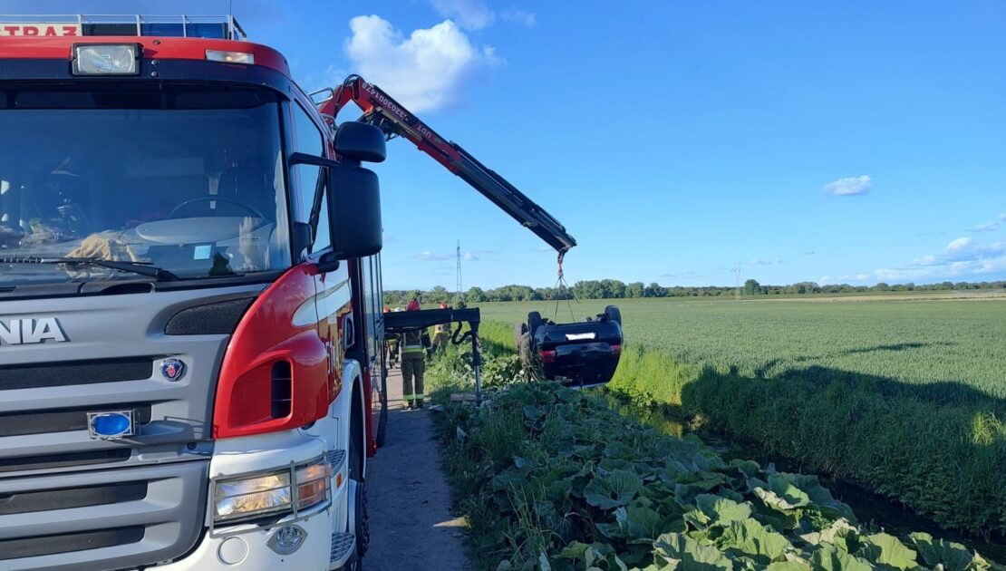 Z lewej strony widać część auta strażackiego, które z tyłu ma wysunięte ramię dźwigu. Do dźwigu podczepione jest auto osobowe, wiszące dachem do dołu. Auto znajduje się nad rowem, w tle pola.