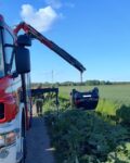 Z lewej strony widać część auta strażackiego, które z tyłu ma wysunięte ramię dźwigu. Do dźwigu podczepione jest auto osobowe, wiszące dachem do dołu. Auto znajduje się nad rowem, w tle pola.