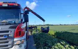 Z lewej strony widać część auta strażackiego, które z tyłu ma wysunięte ramię dźwigu. Do dźwigu podczepione jest auto osobowe, wiszące dachem do dołu. Auto znajduje się nad rowem, w tle pola.