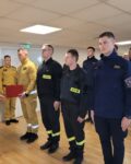 Pododdział strażaków w mundurach stoi na zbiórce w pomieszczeniu. Przed frontem szyku stoi czterech wyróżnionych strażaków