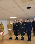 Pododdział strażaków w mundurach stoi na zbiórce w pomieszczeniu. Przed frontem szyku stoi czterech wyróżnionych strażaków