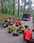 Na leśnej drodze strażący i ratownicy medyczni pochylają się nad osobą poszkodowaną. Z tyłu w tle samochód pożarniczy. Na poboczu stoi skuter.