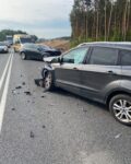Na drodze stoi szary samochód osobowy, po zderzeniu z innym autem osobowym. W tle inne samochody i karetka.
