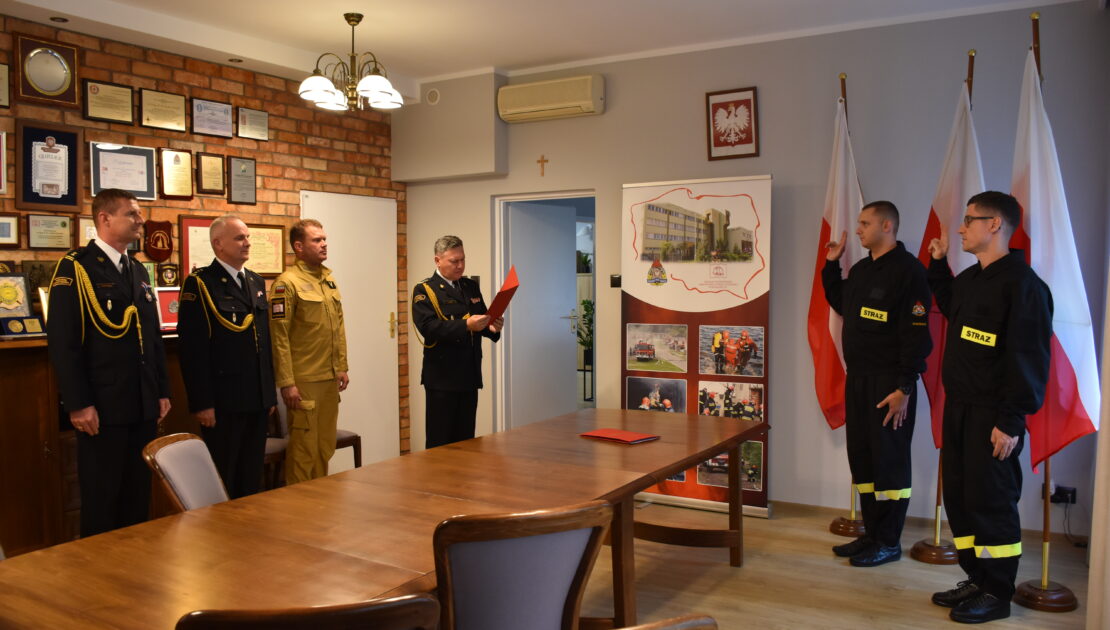Dwóch strażaków w ubraniu koszarowym trzyma podniesioną prawą rękę, stoją po prawej stronie. W tle 3 flagi Polski. Stoją w pomieszczeniu, na ścianie wisi godło Polski. Po lewej stronie stoi 4 strażaków w mundurach wyjściowych