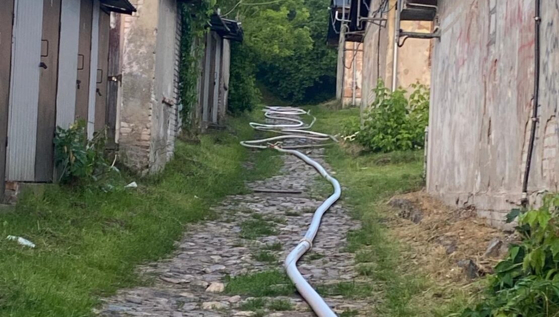 Między budynkami gospodarczymi na ścieżce brukowej leży rozwinięty wąż pożarniczy.