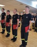 Pododdział strażaków w mundurach stoi na zbiórce w pomieszczeniu. Przed frontem szyku stoi pięciu wyróżnionych strażaków