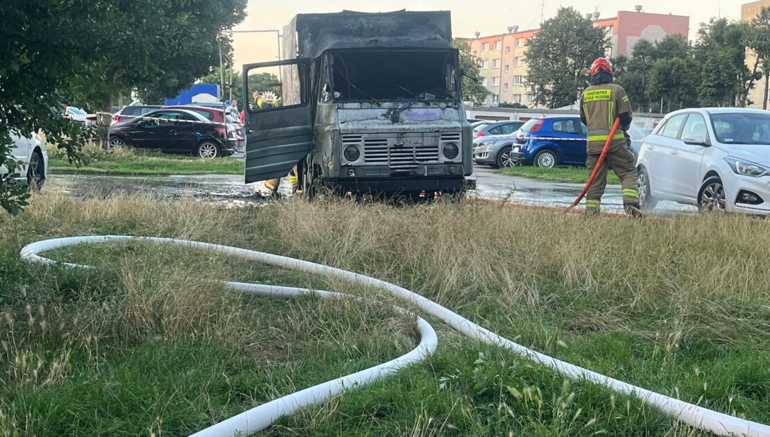 Na parkingu stoi spalony samochód dostawczy. Przed pojazdem widać rozwinięta na trawie linię gaśniczą, którą trzyma strażak i podaje wodę. W tle widać inna zaparkowane auta