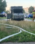 Na parkingu stoi spalony samochód dostawczy. Przed pojazdem widać rozwinięta na trawie linię gaśniczą, którą trzyma strażak i podaje wodę. W tle widać inna zaparkowane auta