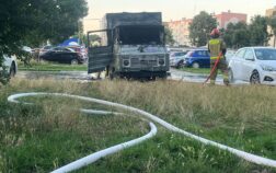 Na parkingu stoi spalony samochód dostawczy. Przed pojazdem widać rozwinięta na trawie linię gaśniczą, którą trzyma strażak i podaje wodę. W tle widać inna zaparkowane auta