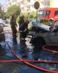 Strażacy stoją przed maską spalonego samochodu osobowego stojącego na parkingu. Na ziemi leżą węże strażackie. W tle samochód gaśniczy i blok mieszkalny
