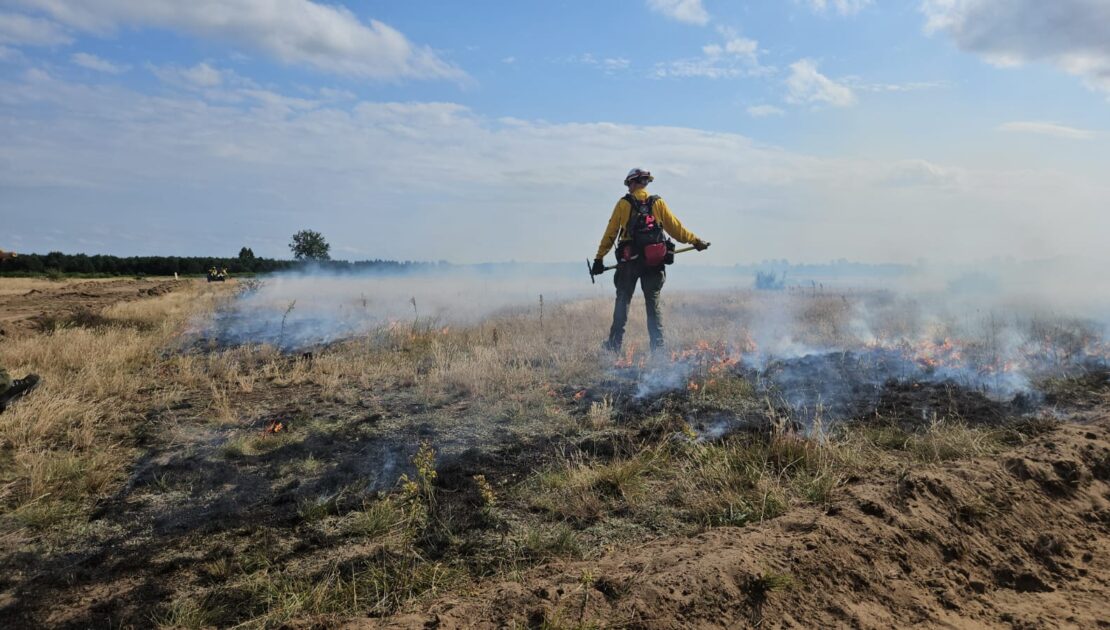 Nad łąką widać unoszący się dym. Na środku tyłem stoi osoba, ma ubrany kask i trzyma coś w rękach