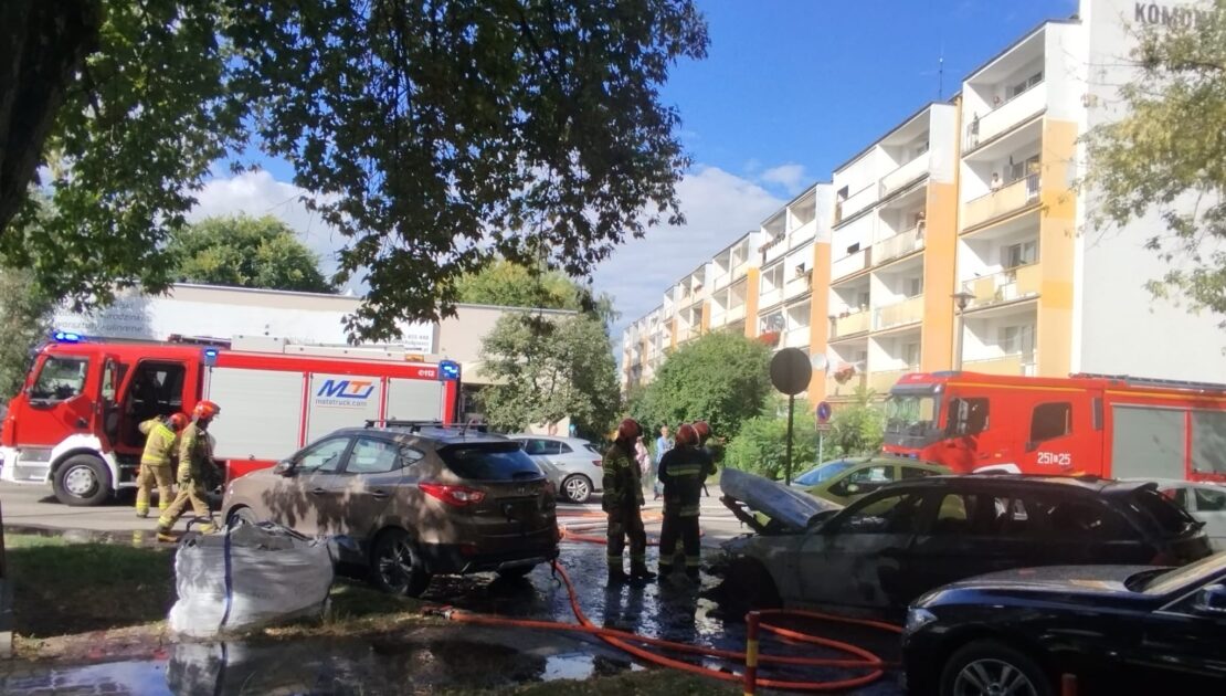 Strażacy stoją przed maską spalonego samochodu osobowego stojącego na parkingu. Obok spalonego auta stoją dwa inne samochody. Na ziemi leżą węże strażackie. W tle dwa samochody gaśnicze i blok mieszkalny