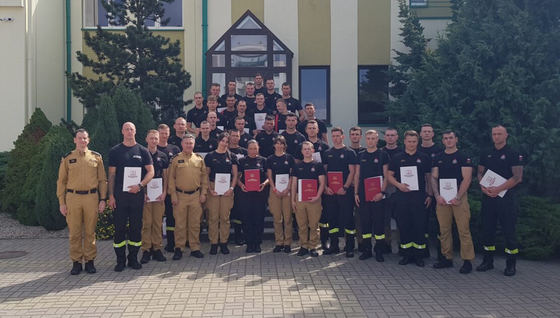 38 strażaków w mundurach koszarowych i służbowych stoją , część z nich trzyma teczki
