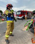 Strażak w mundurze ma ubraną biało-niebieską kamizelkę i mówi coś do radia. W tle samochód strażacki. Po prawej stronie widać kawałek podwozia leżącego auta i podpory zabezpieczające przed przewróceniem się waraku.
