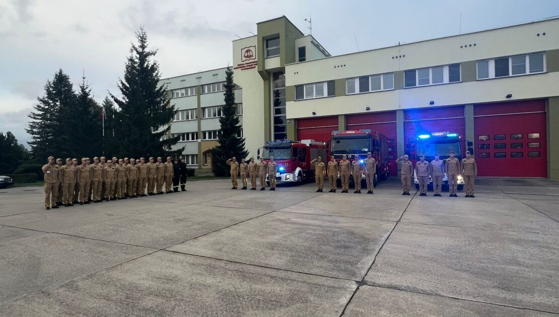 Strażacy stoją przed JRG na zbiórce. W tle samochody i bramy garażowe, auta mają włączone sygnały świetlne.