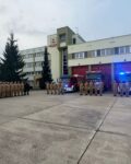 Strażacy stoją przed JRG na zbiórce. W tle samochody i bramy garażowe, auta mają włączone sygnały świetlne.
