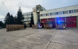 Strażacy stoją przed JRG na zbiórce. W tle samochody i bramy garażowe, auta mają włączone sygnały świetlne.