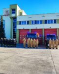 Strażacy stoją przed JRG na zbiórce. W tle samochody i bramy garażowe, auta mają włączone sygnały świetlne.