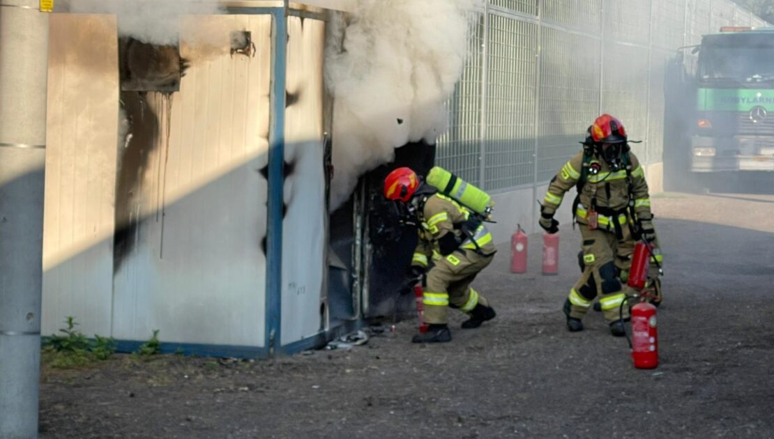 Pożar rozdzielni elektrycznej nad którą unosi się biały dym. Przed wejściem dwóch strażaków z gaśnicą.