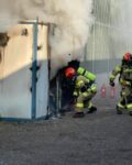 Pożar rozdzielni elektrycznej nad którą unosi się biały dym. Przed wejściem dwóch strażaków z gaśnicą.