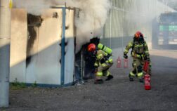 Pożar rozdzielni elektrycznej nad którą unosi się biały dym. Przed wejściem dwóch strażaków z gaśnicą.