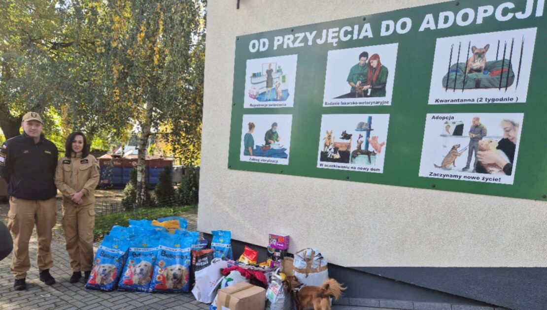 Na placu przed budynkiem leży karma dla zwierząt. Po lewej stronie stoi dwóch strażaków w mundurach służbowych.