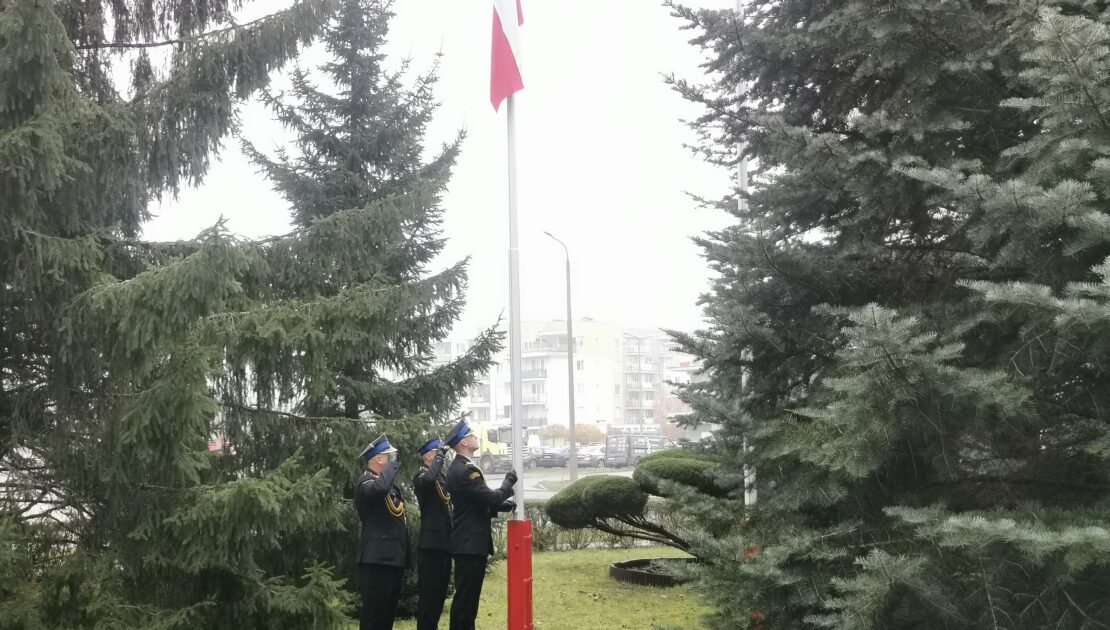 Trzech strażaków w mundurach wyjściowych stoją przy maszcie, na którym wisi flaga Polski