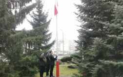 Trzech strażaków w mundurach wyjściowych stoją przy maszcie, na którym wisi flaga Polski