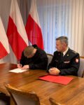 Strażak siedzi przy stole i podpisuje dokumenty, obok siedzi Komendant szkoły