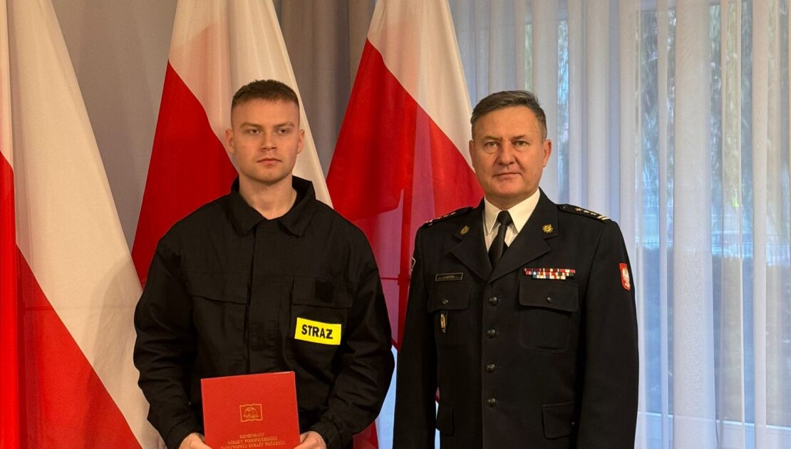 Po lewej stronie stoi strażak i trzyma w ręku czwerwoną teczkę. Po prawej stronie stoi komendant. W tle Flagi Polski.