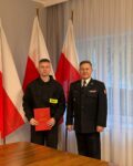 Po lewej stronie stoi strażak i trzyma w ręku czwerwoną teczkę. Po prawej stronie stoi komendant. W tle Flagi Polski.