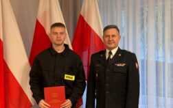 Po lewej stronie stoi strażak i trzyma w ręku czwerwoną teczkę. Po prawej stronie stoi komendant. W tle Flagi Polski.