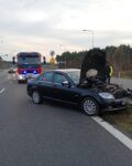 Na jedni stoi uszkodzone auto, ma otwartą maskę. Za nim stoi samochód strażacki.