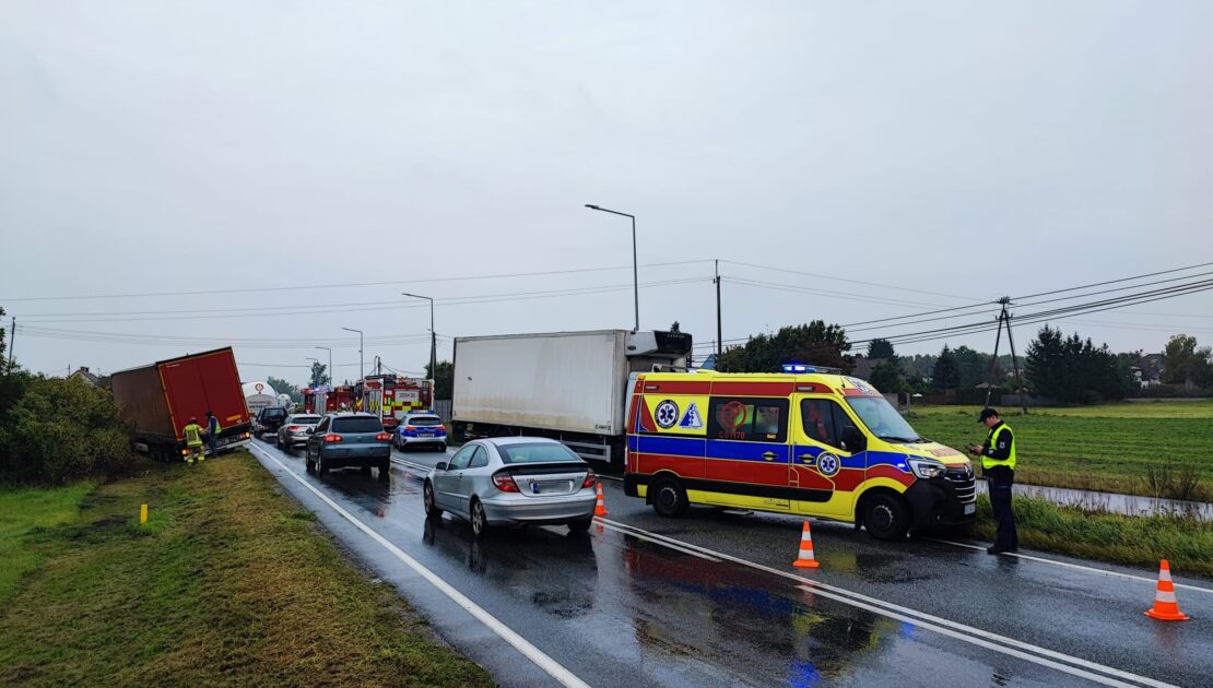 Na drodze stoją dwa uszkodzone pojazdy, oraz ambulans. W tle widać wozy strażackie, Przed uszkodzonym autem po prawej stronie stoi policjant. Przy drugim uszkodzonym aucie stoją strażacy