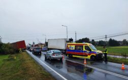 Na drodze stoją dwa uszkodzone pojazdy, oraz ambulans. W tle widać wozy strażackie, Przed uszkodzonym autem po prawej stronie stoi policjant. Przy drugim uszkodzonym aucie stoją strażacy