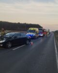 Na jedni stoi uszkodzone auto z wgniecioną maską. Za nim stoi ambulans i wóz strażacki. W tle widać stojące na jedni auta. Obok uszkodzonego pojazdu jest strażak.