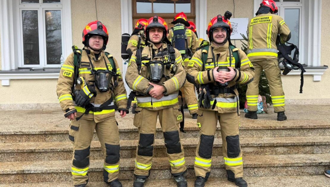 Trzech strażaków stoi na schodach. Są ubraniu w ubrania specjalne a na głowie mają hełmy.