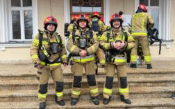 Trzech strażaków stoi na schodach. Są ubraniu w ubrania specjalne a na głowie mają hełmy.